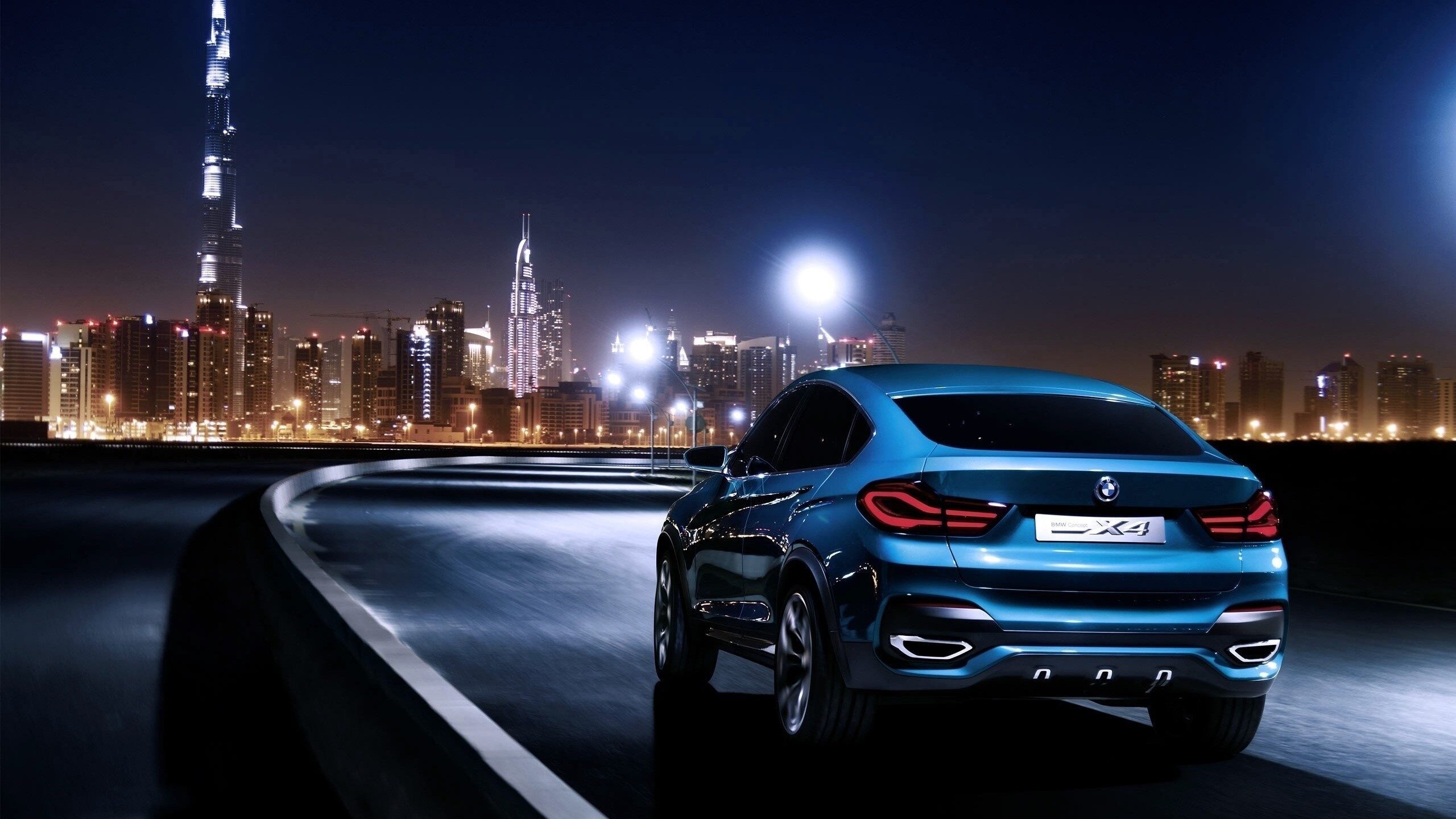 Rear view of a blue BMW X4 driving at night with a city skyline, showcasing a luxury vehicle that can receive expert auto body repair at Westside Valley Auto Body